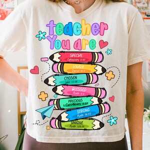 May include: Cream-colored t-shirt with a colorful graphic design. The design features the words "teacher You are" above stacked pencils, each with a different color and a positive adjective like "Special" and "Unique".