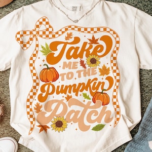 Könnte beinhalten: Cremefarbenes T-Shirt mit Retro-Design. Die Grafik zeigt den Text "Take Me To The Pumpkin Patch" in Orange, mit Kürbissen, Sonnenblumen und Herbstblättern. Ein kariertes Band umrahmt das Design.