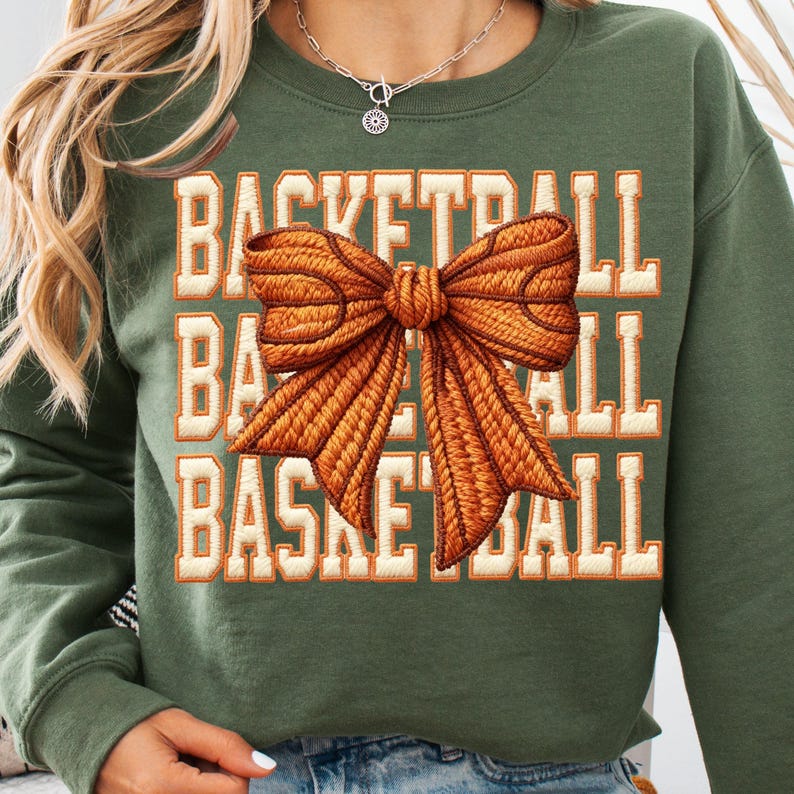 May include: Olive green sweatshirt with the word "BASKETBALL" in a stacked design. A large, textured orange bow is centered on the front. The text is in a block letter style with an orange outline.