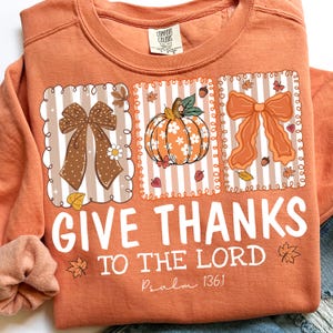 May include: Orange sweatshirt with a graphic design featuring bows, a pumpkin, and fall leaves. The text "GIVE THANKS TO THE LORD" is printed in white, with "Psalm 136:1" below. A Comfort Colors tag is visible.