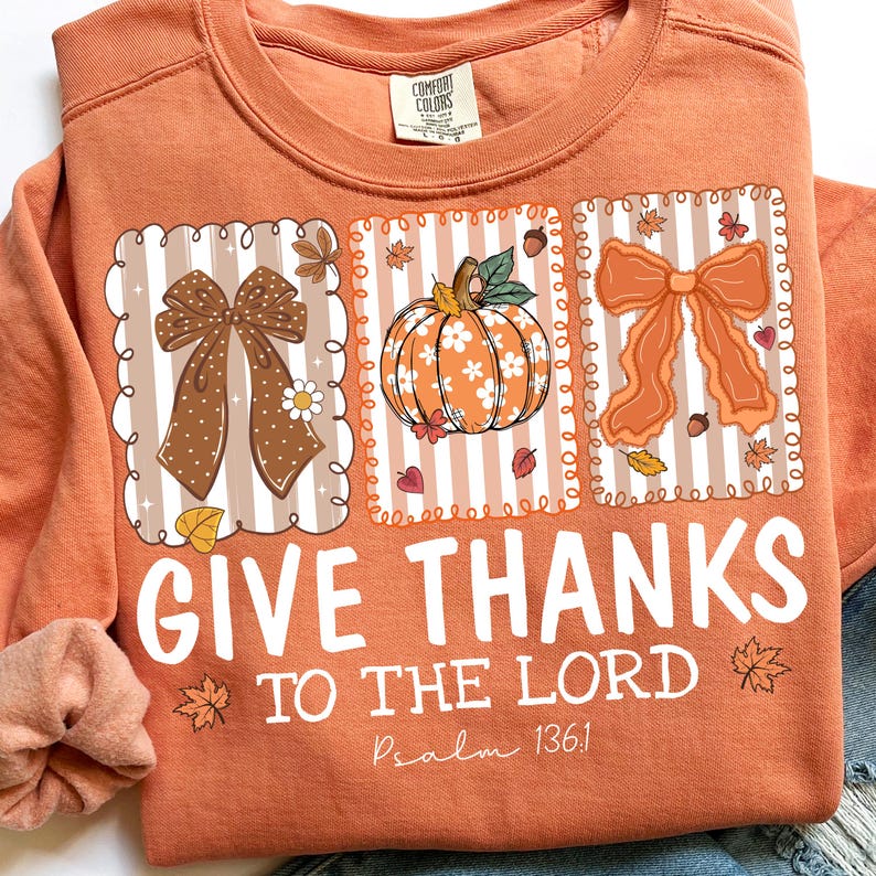 May include: Orange sweatshirt with a graphic design featuring bows, a pumpkin, and fall leaves. The text "GIVE THANKS TO THE LORD" is printed in white, with "Psalm 136:1" below. A Comfort Colors tag is visible.