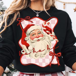 May include: Black sweatshirt featuring a Santa Claus graphic with a red hat, white beard, and candy cane. The design includes the words "ho ho ho" in a cursive font, set against a pink and white checkered background. Red bows accent the design.