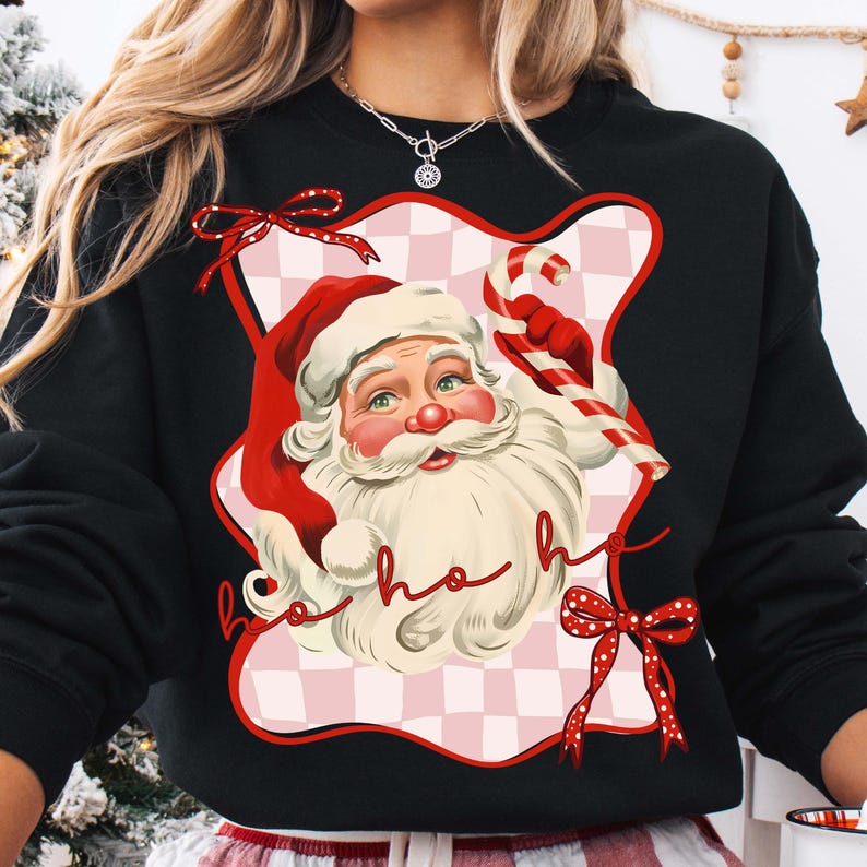 May include: Black sweatshirt featuring a Santa Claus graphic with a red hat, white beard, and candy cane. The design includes the words "ho ho ho" in a cursive font, set against a pink and white checkered background. Red bows accent the design.