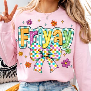 Peut inclure: Sweat-shirt rose avec un motif color&eacute; du mot "Friyay" avec un n&oelig;ud &agrave; carreaux. Le motif est entour&eacute; de petites fleurs et d'&eacute;toiles.