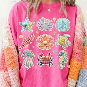 Pode incluir: Uma t-shirt rosa com bordados de criaturas marinhas coloridas, incluindo uma estrela-do-mar, uma concha, um caranguejo e um cavalo-marinho. O design apresenta vários padrões e cores, criando uma estética de verão caprichosa. Gola redonda e mangas curtas.