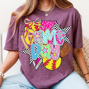 May include: Purple t-shirt with a colorful graphic design featuring the words "Game Day" with a baseball, bat, and glove.