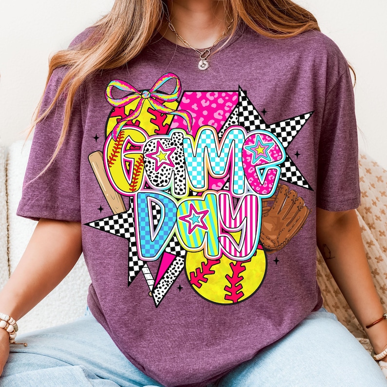 May include: Purple t-shirt with a colorful graphic design featuring the words "Game Day" with a baseball, bat, and glove.