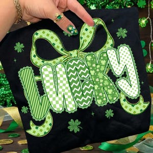 Puede incluir: Camiseta negra con la palabra "LUCKY" en letras decorativas, adornada con un lazo verde y motivos de tr&eacute;boles. Las letras est&aacute;n decoradas con rayas, chevrones y lunares. Un dise&ntilde;o festivo para el D&iacute;a de San Patricio.