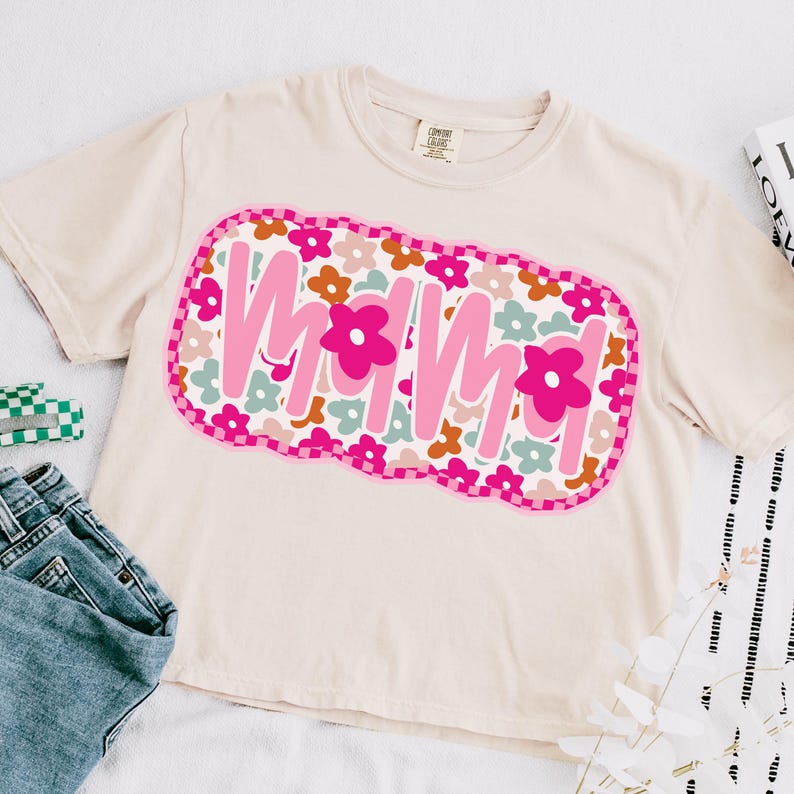 May include: A light beige t-shirt with a pink, green, and orange graphic that says "MOMMA" surrounded by flowers.