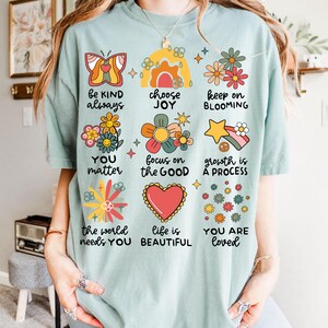 May include: A light blue t-shirt with a variety of colorful illustrations and text. The text includes phrases like "be kind always", "choose joy", "keep on blooming", "you matter", "focus on the good", "growth is a process", "the world needs you", "life is beautiful", and "you are loved".