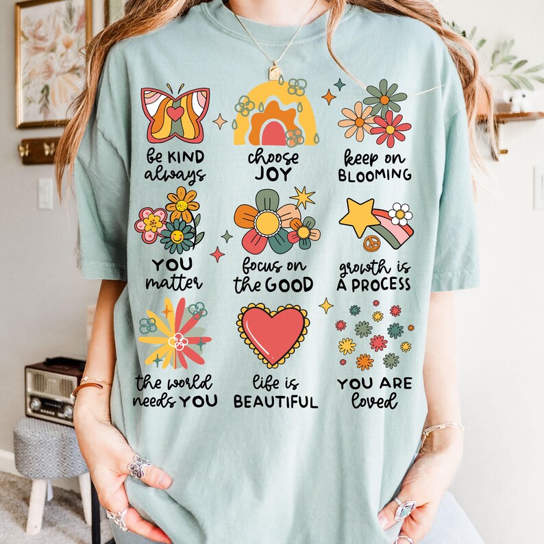 May include: A light blue t-shirt with a variety of colorful illustrations and text. The text includes phrases like "be kind always", "choose joy", "keep on blooming", "you matter", "focus on the good", "growth is a process", "the world needs you", "life is beautiful", and "you are loved".