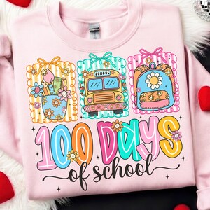 K&ouml;nnte beinhalten: Hellrosa Sweatshirt mit einem farbenfrohen Grafikdesign zum 100. Schultag. Das Design zeigt einen Schulbus, einen Rucksack und einen Stifthalter, alle mit Blumen verziert. Der Text "100 Days of School" ist deutlich sichtbar.