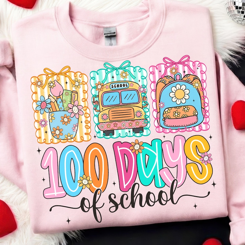 K&ouml;nnte beinhalten: Hellrosa Sweatshirt mit einem farbenfrohen Grafikdesign zum 100. Schultag. Das Design zeigt einen Schulbus, einen Rucksack und einen Stifthalter, alle mit Blumen verziert. Der Text "100 Days of School" ist deutlich sichtbar.