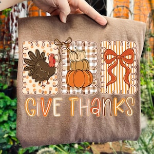 May include: Brown t-shirt with a Thanksgiving-themed design. The design includes a turkey, stacked pumpkins, and a large bow, all within decorative frames. The words "GIVE THANKS" are written in a fall-themed color palette.