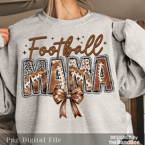 May include: Grey sweatshirt with the text "Football Mama" in brown and white lettering. The letters are decorated with brown footballs and leopard print. A brown bow is below the text.