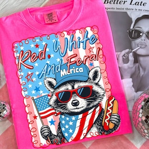 May include: Bright pink t-shirt with a graphic of a raccoon wearing sunglasses, a bandana, and a baseball cap, holding an American flag and a hot dog. The text reads "Red, White, and Feral, 'Murica" in red and blue.