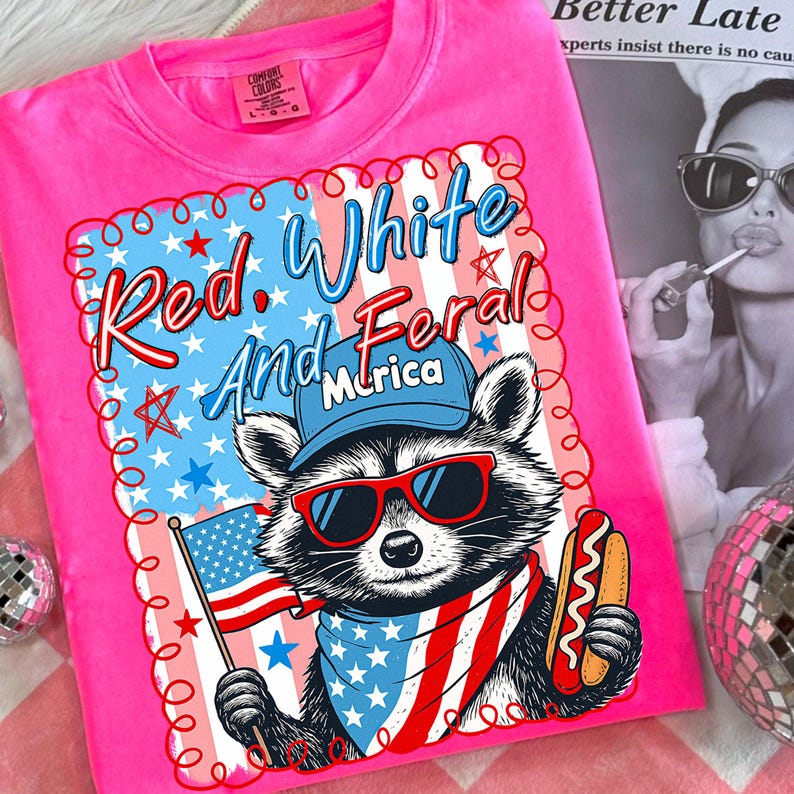 May include: Bright pink t-shirt with a graphic of a raccoon wearing sunglasses, a bandana, and a baseball cap, holding an American flag and a hot dog. The text reads "Red, White, and Feral, 'Murica" in red and blue.