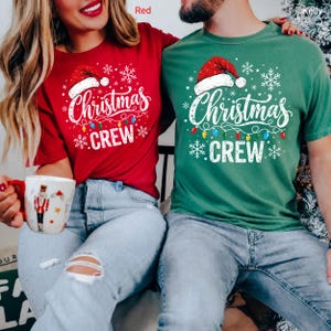 Puede incluir: Una camiseta roja y una camiseta verde kelly, cada una con un gráfico blanco de un gorro de Papá Noel, copos de nieve y el texto "Christmas CREW" sobre una cadena de luces navideñas de colores. Se ve una taza blanca con un cascanueces.