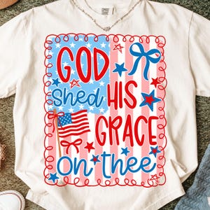 May include: White t-shirt with a patriotic design. The design features the text "GOD SHED HIS GRACE ON THEE" in red and blue, with an American flag and stars. The shirt is made of a soft material.