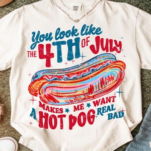 May include: Cream-colored t-shirt with a colorful graphic of a hot dog and text. The text reads "You look like the 4th of July makes me want a hot dog real bad." The design features red, blue, and orange hues.