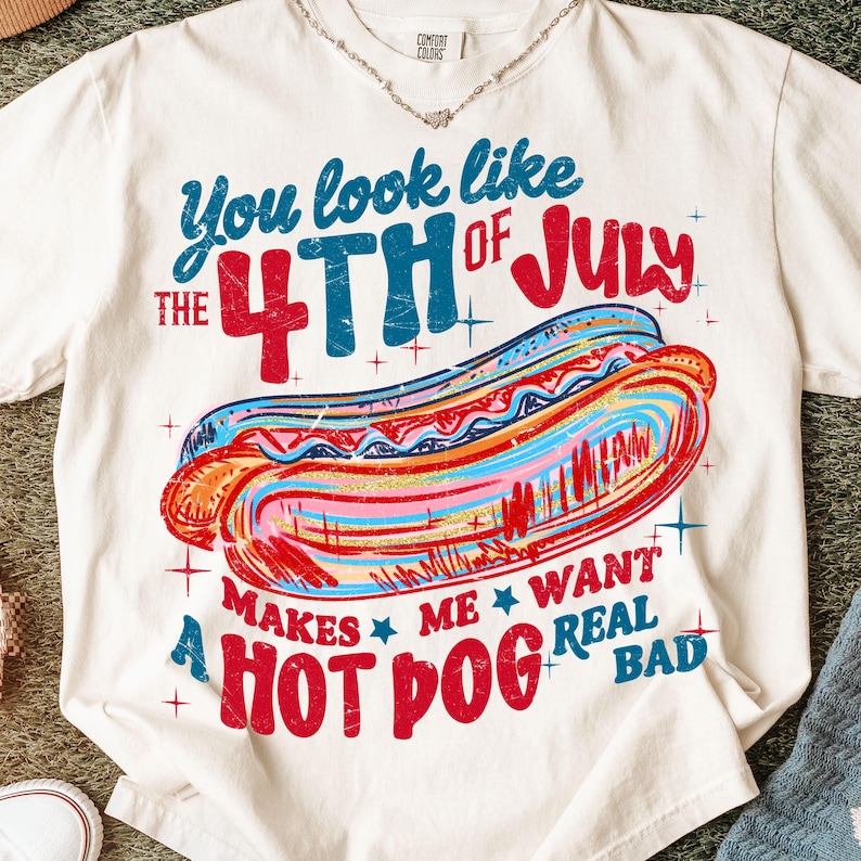 May include: Cream-colored t-shirt with a colorful graphic of a hot dog and text. The text reads "You look like the 4th of July makes me want a hot dog real bad." The design features red, blue, and orange hues.
