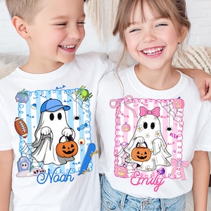 Puede incluir: Dos niños con camisetas blancas con diseños de fantasmas de Halloween. Una camiseta tiene un marco azul con un fantasma con gorra de béisbol, sosteniendo una calabaza. La otra camiseta tiene un marco rosa con un fantasma con un lazo, también sosteniendo una calabaza.
