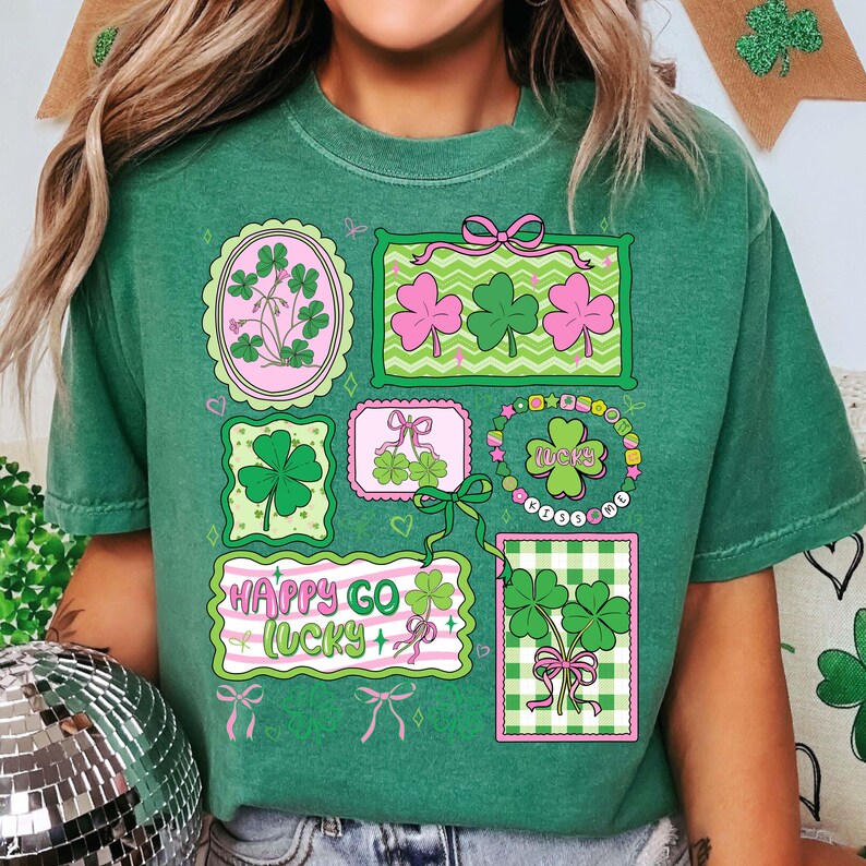Op de afbeelding: Groen t-shirt met diverse klaverontwerpen in roze en groene kaders. Het shirt bevat de tekst "Happy Go Lucky" en is versierd met strikken en linten, perfect voor St. Patrick's Day.