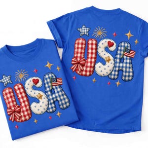 K&ouml;nnte beinhalten: Zwei blaue T-Shirts mit dem Wort "USA" auf der Vorderseite. Die Buchstaben sind in rot-wei&szlig;em Vichy, Wei&szlig; mit blauen Punkten und blauem Vichy gehalten. Jedes Shirt hat Stern- und Feuerwerksstickereien.