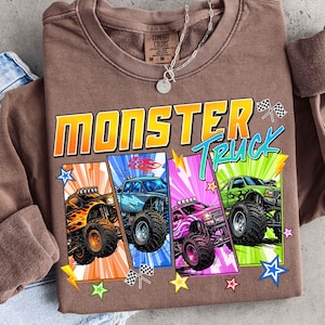 May include: Brown crewneck sweatshirt with a colorful graphic of four monster trucks. The design includes the words "Monster Truck" in a bold, outlined font, along with stars and checkered flags.