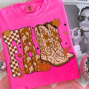 May include: Vibrant pink t-shirt with the word "FA" in a brown and tan checkered and cactus design. The letter "R" features cowboy boots with floral and leopard print. A pumpkin, horseshoe, and autumn leaves complete the design.