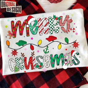 May include: White t-shirt with the words "MERRY CRUISMAS" in colorful, festive lettering. The design includes Christmas-themed elements like a Santa hat, gingerbread man, cruise ships, and string lights, set against a red and black plaid background.