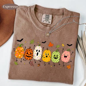 May include: An espresso-colored t-shirt featuring a graphic print of six cartoon pumpkins and a ghost with various facial expressions and floral patterns, surrounded by small bats, leaves, and stars. The shirt has a crew neckline and a brand label.