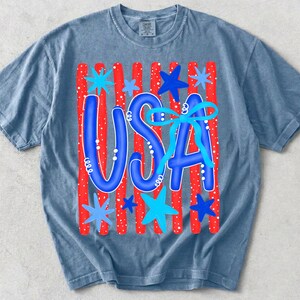 May include: A blue t-shirt with a graphic design featuring the word "USA" in blue, decorated with a blue bow and stars. The background has red stripes with white dots, creating a patriotic theme.