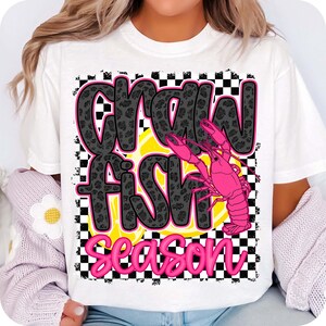 May include: White t-shirt with a black and white checkered background. The text "Crawfish Season" is written in black with a leopard print pattern. A pink crawfish is on top of a yellow lemon slice.
