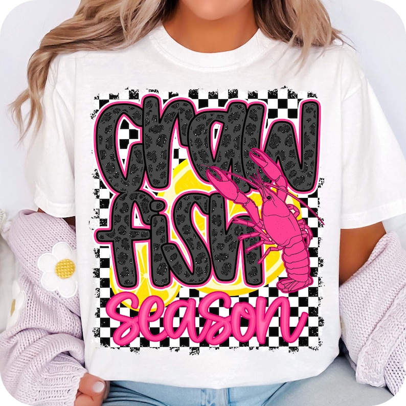 May include: White t-shirt with a black and white checkered background. The text "Crawfish Season" is written in black with a leopard print pattern. A pink crawfish is on top of a yellow lemon slice.