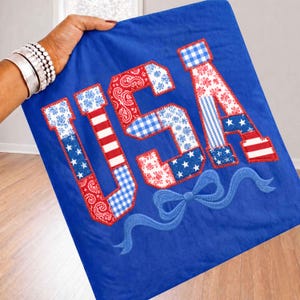May include: A blue textile featuring the letters "USA" in a patchwork design. The letters are made up of red, white, and blue fabrics with patterns like stars, stripes, and paisley. A blue embroidered bow is at the bottom.