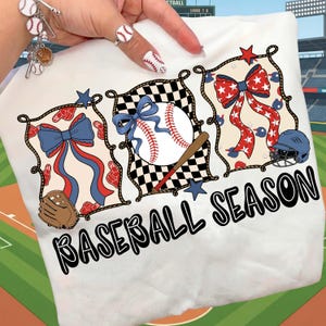 Puede incluir: Camiseta blanca con un diseño temático de béisbol. El diseño incluye una pelota de béisbol, un bate y un lazo, con las palabras "BASEBALL SEASON" en negro. El diseño incorpora los colores rojo, blanco y azul.