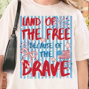 以下が含まれることがあります： 愛国的なデザインの白いTシャツ。「LAND OF THE FREE BECAUSE OF THE BRAVE」のテキストが赤色で、青と白のストライプの背景、花火、星、アメリカ国旗が描かれています。