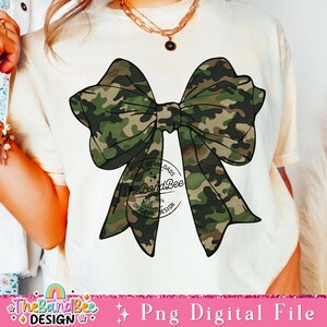 May include: A white t-shirt with a camouflage bow design. The bow is green, brown, and black. The shirt also has a small logo that says "The Band Bee" with the words "Digital Downloads, Est. 2014, Original Design".