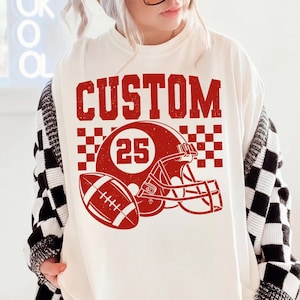 May include: An off-white crewneck t-shirt featuring a red graphic print. The design includes the word CUSTOM in a distressed font above a football helmet with the number 25, a football, and a checkered pattern background element.