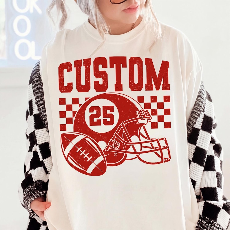 May include: An off-white crewneck t-shirt featuring a red graphic print. The design includes the word CUSTOM in a distressed font above a football helmet with the number 25, a football, and a checkered pattern background element.