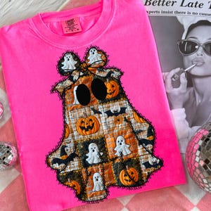 May include: A bright pink Comfort Colors sweatshirt featuring a patchwork ghost design. The ghost is made of orange, black, and white squares with Halloween-themed images. The ghost has large black eyes and is outlined with black stitching.