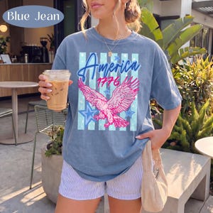 Puede incluir: Camiseta azul vaquera con un &aacute;guila rosa, estrellas azules y la inscripci&oacute;n "America 1776". La camiseta se combina con pantalones cortos a rayas. La imagen est&aacute; tomada al aire libre, con vegetaci&oacute;n de fondo.