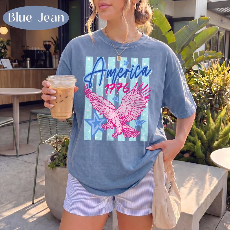 Puede incluir: Camiseta azul vaquera con un &aacute;guila rosa, estrellas azules y la inscripci&oacute;n "America 1776". La camiseta se combina con pantalones cortos a rayas. La imagen est&aacute; tomada al aire libre, con vegetaci&oacute;n de fondo.
