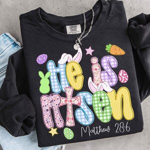 May include: Black Comfort Colors sweatshirt with the words "He is Risen" in colorful, patterned letters. The design includes Easter-themed graphics like a carrot, eggs, and bunny ears. The text "Matthew 28:6" is printed below the main text.