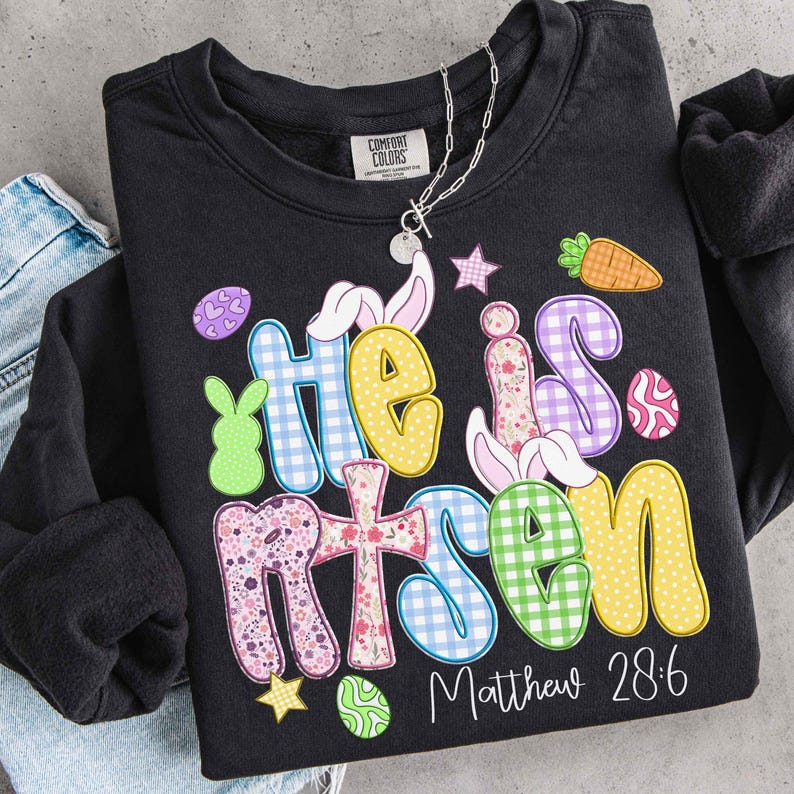 May include: Black Comfort Colors sweatshirt with the words "He is Risen" in colorful, patterned letters. The design includes Easter-themed graphics like a carrot, eggs, and bunny ears. The text "Matthew 28:6" is printed below the main text.