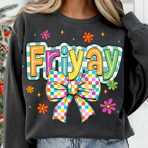 Peut inclure: Un sweat-shirt gris fonc&eacute; avec un motif graphique color&eacute; repr&eacute;sentant le mot "Friyay" avec un n&oelig;ud &agrave; carreaux. Le graphique est de style r&eacute;tro avec des couleurs vives et un motif &agrave; carreaux.