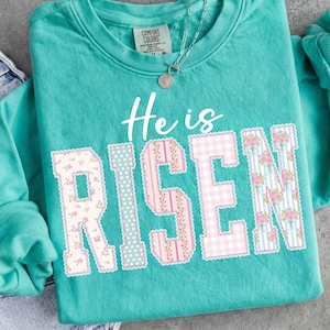 May include: Teal sweatshirt with the phrase "He is RISEN" in white and patchwork letters. The letters have floral, polka dot, and gingham patterns in pink and white. A silver necklace with a pendant is visible.