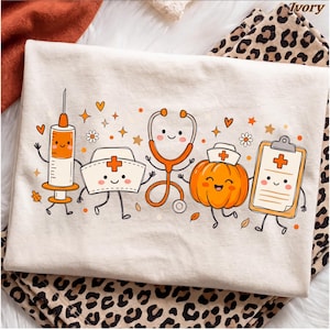 May include: An ivory t-shirt featuring a graphic print of cartoon medical items including a syringe, a nurse cap, a stethoscope, a pumpkin wearing a nurse cap, and a clipboard, all with smiling faces, surrounded by orange hearts, stars, and leaves.