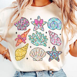 May include: A white t-shirt with a floral pattern of seashells, starfish, and coral. The design is in shades of pink, blue, yellow, and green.